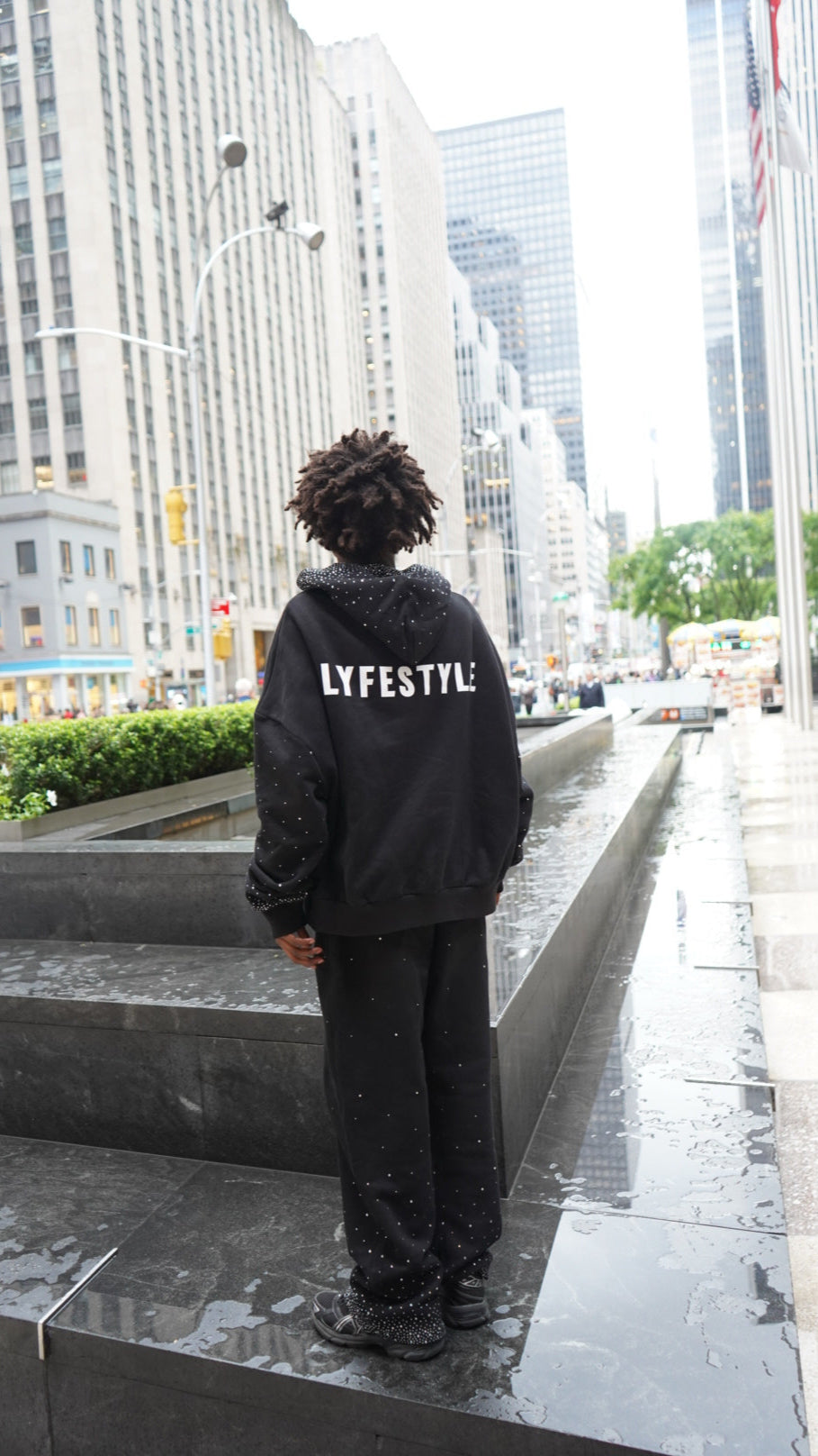Person wearing a black hoodie with text, standing on a concrete ledge with a cityscape background