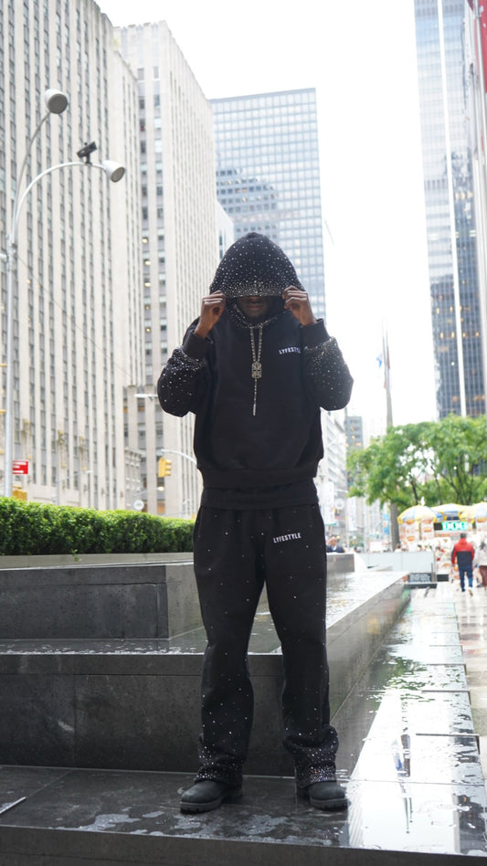 Person wearing a black coat and hat in an urban setting with buildings and trees in the background.